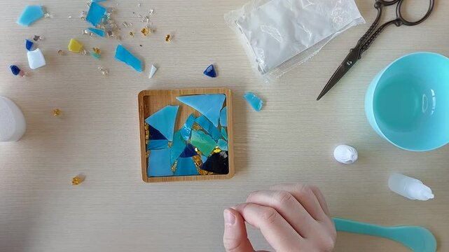 Finishing mosaic design for handmade coaster by placing final colorful glass fragment into adhesive-covered wooden mold