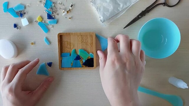 Continuing mosaic layout by placing more glass fragments into wooden mold for coaster creation using handmade gypsum and colorful materials
