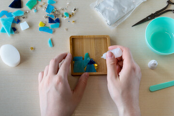 Placing final piece of faux glass into silicone mold during crafting of handmade gypsum mosaic coaster with colorful fragments and table setup, step 1