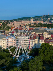 Lush green trees are around the giant ferris wheel. Buda royal castle is on the background.