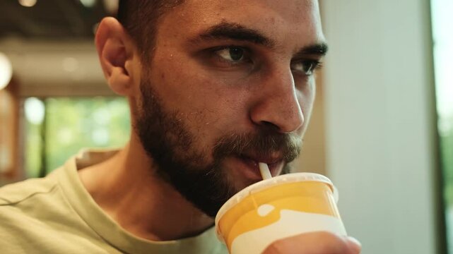 Young man drinking soda from a paper cup with a straw in a fast food restaurant, seated by a window with natural light. Horizontal 4k footage