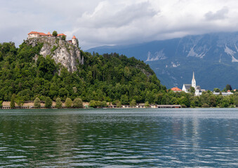A panoramic view of the iconic landmarks surrounding Lake Bled, Slovenia. On the left, Bled Castle majestically perches atop a dramatic rocky cliff