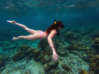 A young woman swims gracefully underwater, exploring colorful coral reefs. Sunlight filters through the water, creating a vibrant, serene atmosphere in a tropical location.