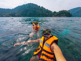 Two individuals wearing bright life vests are snorkeling in clear waters. They are exploring a vibrant underwater world while surrounded by lush green mountain scenery.