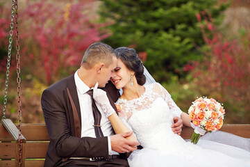A bride and groom are sitting on a bench, holding hands and smiling
