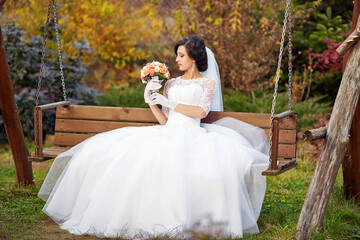 A woman in a white wedding dress sits on a swing, holding a bouquet of flowers