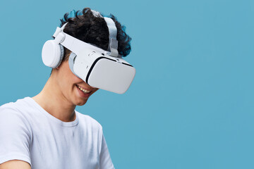 young man wearing virtual reality headset and white headphones smiling against blue background,...