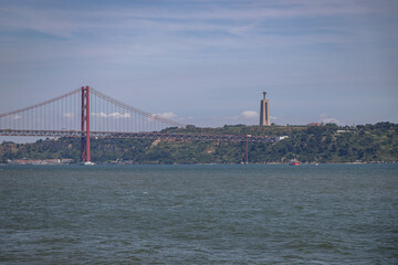 View of the 25 de Abril Bridge and the Cristo Rei monument across the Tagus River, photographed from Lisbon, Portugal, on June 13, 2025, on a clear day with calm water.