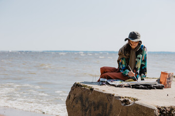 a young artist girl is sitting on the beach painting a sunny day with strong winds and waves on the...