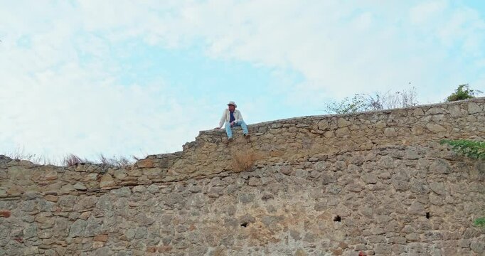 A young adult man wearing a hat and glasses sits on a medieval wild stone wall against a blue sky
