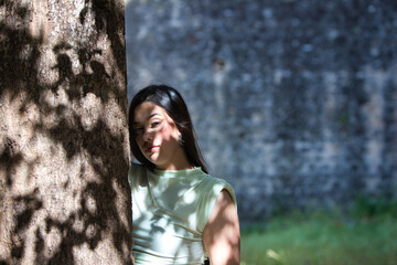 A beautiful Latin teenager with long dark hair rests her hands and face on a tree trunk in the...