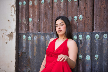 A pretty teenage girl with long dark hair, dressed in red and wearing high heels, sitting on the steps of a church and leaning against the wooden door.