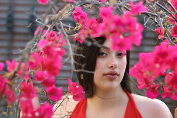 Portrait of a pretty teenage girl with dark hair, dressed in red and surrounded by bougainvillea flowers in a street park.