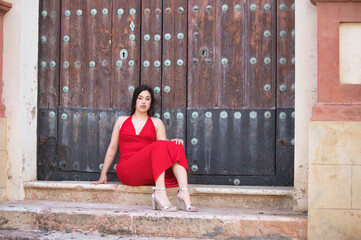 A pretty teenage girl with long dark hair, dressed in red and wearing high heels, sitting on the steps of a church and leaning against the wooden door.