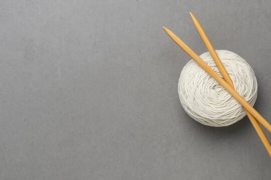 Two wooden knitting needles resting in the corner with ample space for text, alongside a wire cotton bobbin on a gray background, representing hobby supplies and the knitting craft.