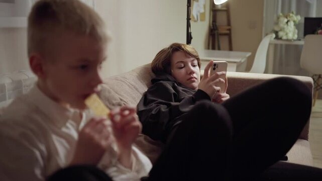 Young mother and her son wearing hearing aids share popcorn while laughing at a TV show. Warm living room scene showcasing joyful family bonding and inclusive representation. Focus on normalizing assi