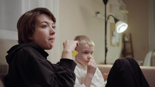 Young mother and her son wearing hearing aids share popcorn while laughing at a TV show. Warm living room scene showcasing joyful family bonding and inclusive representation. Focus on normalizing assi