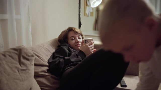 Young mother and her son wearing hearing aids share popcorn while laughing at a TV show. Warm living room scene showcasing joyful family bonding and inclusive representation. Focus on normalizing assi