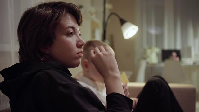 Young mother and her son wearing hearing aids share popcorn while laughing at a TV show. Warm living room scene showcasing joyful family bonding and inclusive representation. Focus on normalizing assi