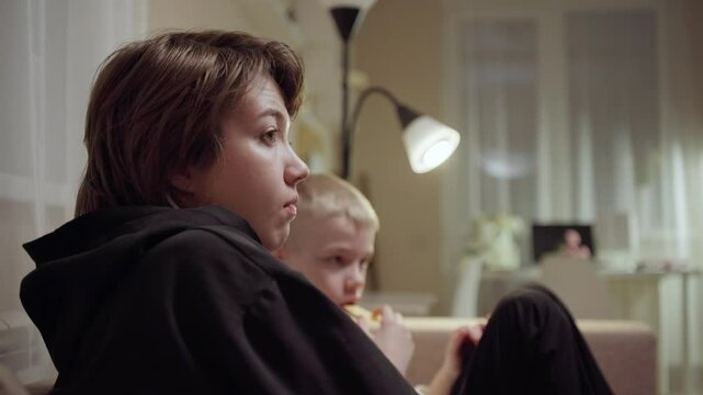 Young mother and her son wearing hearing aids share popcorn while laughing at a TV show. Warm living room scene showcasing joyful family bonding and inclusive representation. Focus on normalizing assi