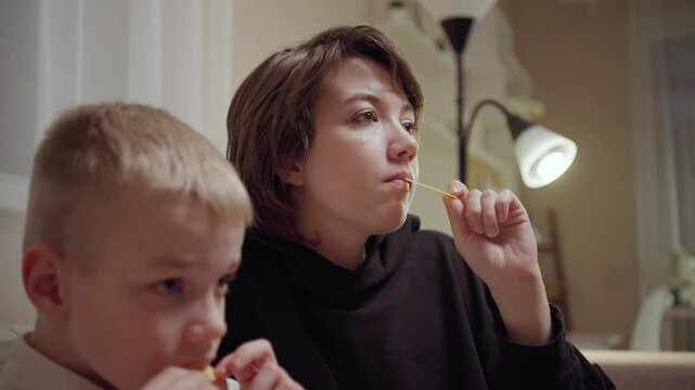 Young mother and her son wearing hearing aids share popcorn while laughing at a TV show. Warm living room scene showcasing joyful family bonding and inclusive representation. Focus on normalizing assi