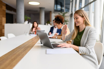 Smiling businesswoman waving at laptop during video call while colleagues working in the background in meeting room