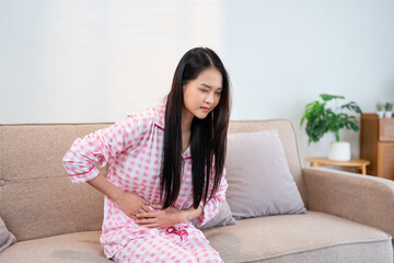 young asian woman in pajamas sitting on couch in living room at home, touching belly, suffering...