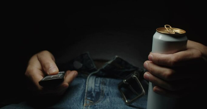 Modern Adult man in jeans with unbuttoned belt and T-shirt sits with metal can with drink and TV remote control. Close-up on stomach and hands