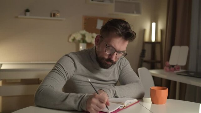 Bearded 30-year-old man sits in the living room in the evening, reading his daily planner and writing down ideas and thoughts. A natural, unaltered moment of focus and reflection in a cozy atmosphere