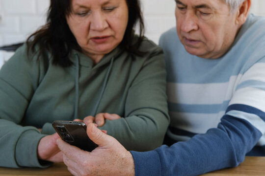 Old couple using a phone together while relaxing in the kitchen at home. Elderly man and woman spending leisure time browsing online with a smartphone. Happy senior married couple typing, learning - Powered by Adobe