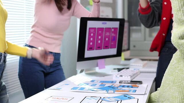 Ui/ux designers clapping hands and high-fiving each other, celebrating successful mobile application development around a table with computer and paper prototypes.