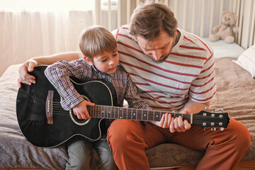 Handsome dad teaches his cute son to play the guitar sitting on sofa in the living room at home....