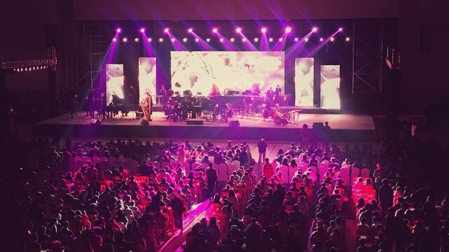 A purple-lit stage hosts a live concert. Musicians play instruments, screens show visuals, and a packed audience fills the venue.
