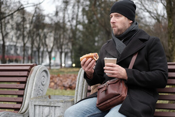 Pensive hipster drinking coffee sitting on a bench in the urban park. Handsome man wearing black...