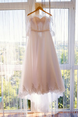 Dress hangs by sunny window. A stunning white wedding dress by a bright window captures sunlight...