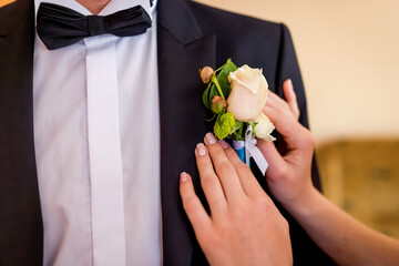 Groom preps for the wedding. A woman attaches a small white rose boutonniere to a man's tuxedo just...