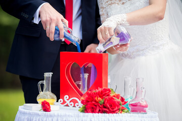 Couple unites with colored sand. A bride and groom pour colorful sand into a heart-shaped container during a unity ceremony at an outdoor wedding.