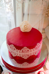 Red celebration cake. A vibrant red cake with elegant lace detailing and a flower on top sits on a decorative stand indoors.