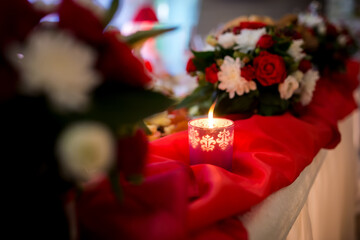 Decorative candle surrounded by flowers. A beautiful candle glows gently on a red cloth, surrounded by vibrant flowers at a festive gathering.