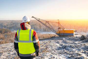 Worker observing winter coal mining operation with excavator at sunrise