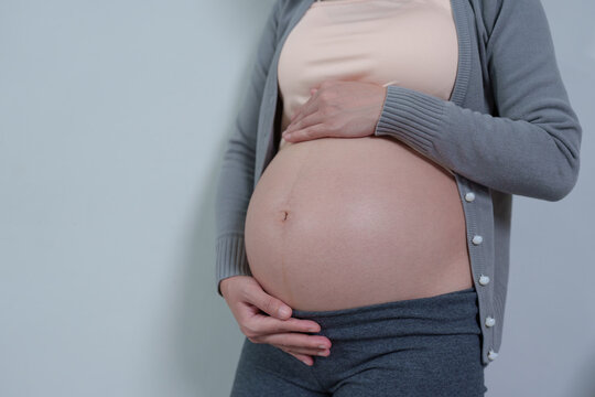 Close-up of a pregnant woman gently cradling her bare belly, wearing a soft gray cardigan and pants, symbolizing motherhood and prenatal care.