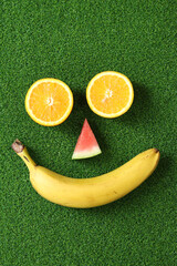 Fruit face on green grass, top view. Two halves of orange, a piece of watermelon and a banana make...