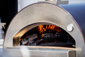 flames and burning wood inside stainless steel outdoor pizza oven with open door during cooking...