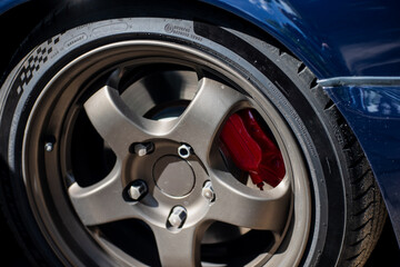 close up of custom alloy wheel and red brake caliper on a blue sports car parked on pavement during...