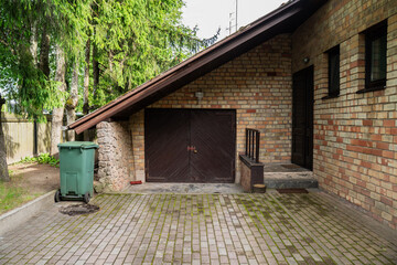 brick house garage exterior with sloped roof, brown metal doors, paved driveway, green trash bin...