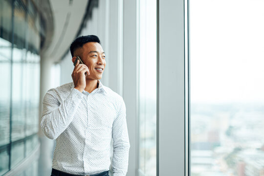 A man in a white shirt smiles while talking on a phone near a large window in a modern office building.