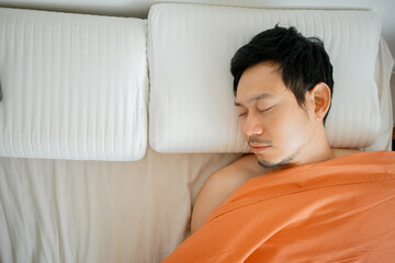 Asian man sleeping in bed under orange blanket on white bed.