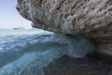 Powerful wave hitting under limestone overhang with sailboat on bright turquoise sea