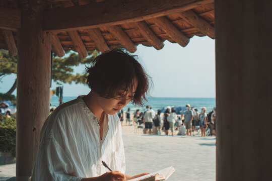 Journaling moment during an art break, teen sketching or writing in a notebook under a shaded pavilion on Gyeongpo Beach, distant sound of waves, personal reflection and cultural exchange.