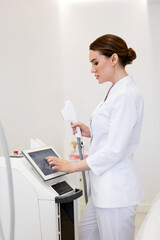 Woman cosmetologist in white coat adjusting laser device before non-surgical skin rejuvenation...